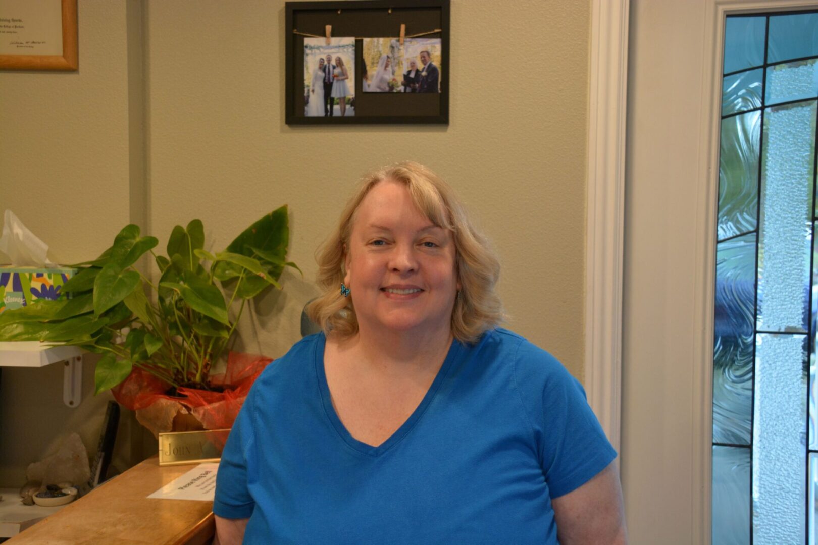 A smiling woman in a blue shirt poses indoors with plants and framed photos behind her.